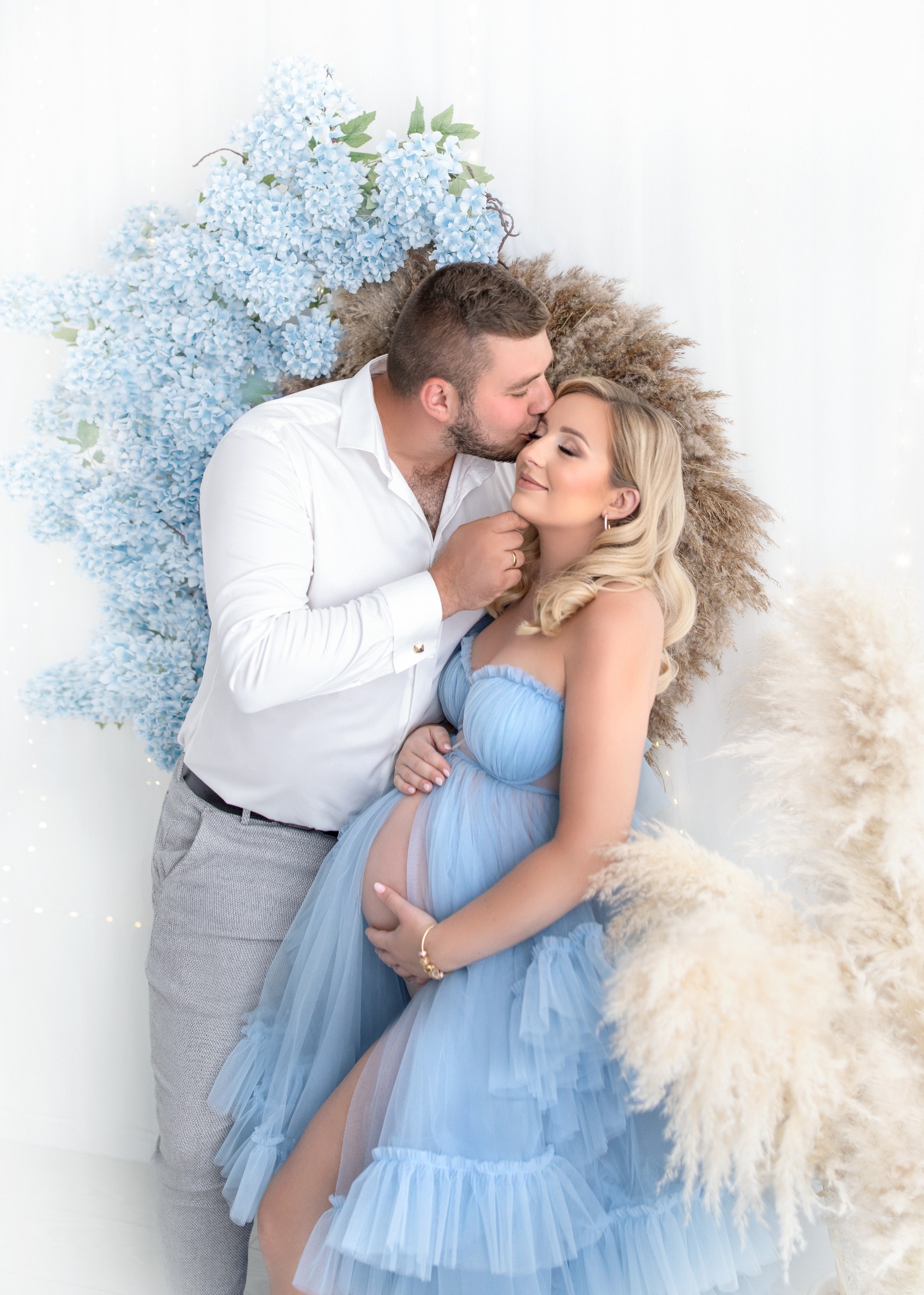 Przyszły tata całuje w czoło uśmiechniętą, ciężarną kobietę w błękitnej sukni na tle białej ściany z dekoracją z błękitnych kwiatów i pampasów