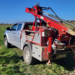 Srebrny pickup z czerwoną wiertnicą Staltehnika na zielonym polu pod błękitnym niebem. Wiertło w ziemi, widoczne panele sterowania i hydraulika.