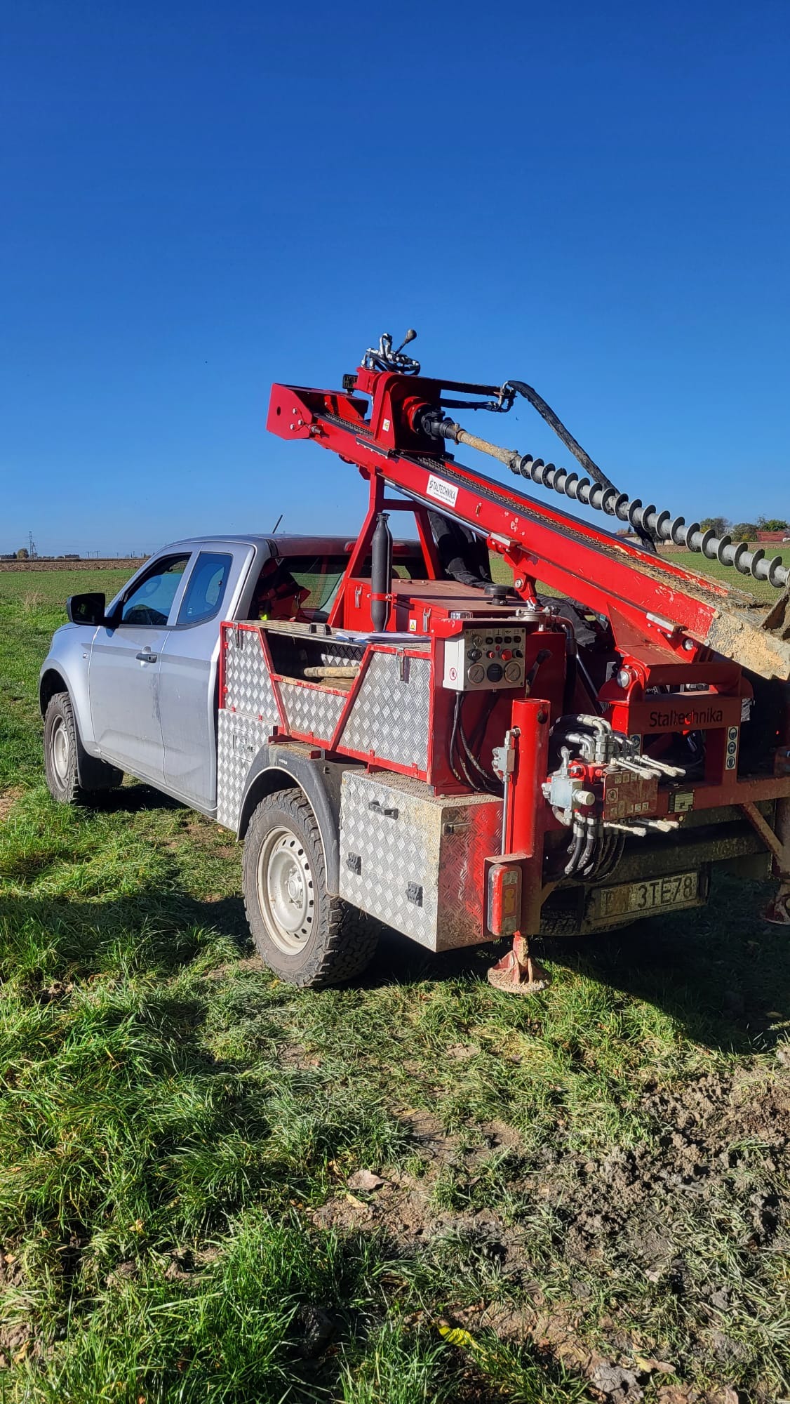 Srebrny pickup z czerwoną wiertnicą Staltehnika na zielonym polu pod błękitnym niebem. Wiertło w ziemi, widoczne panele sterowania i hydraulika.