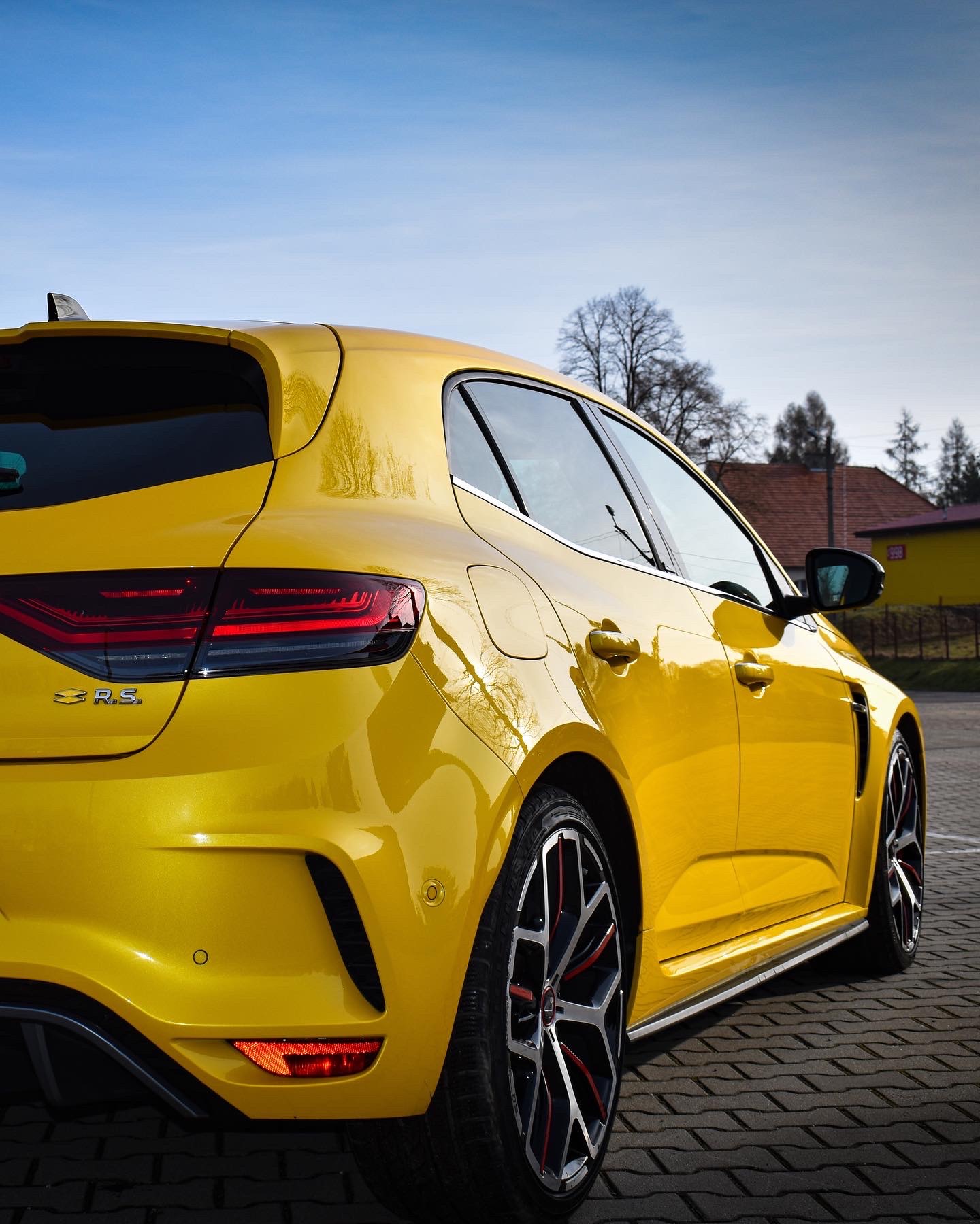 Żółty samochód Renault Megane R.S. zaparkowany na kostce brukowej, widok z tyłu i boku, z widocznymi felgami z czerwonymi akcentami i odbiciami światła na lakierze.