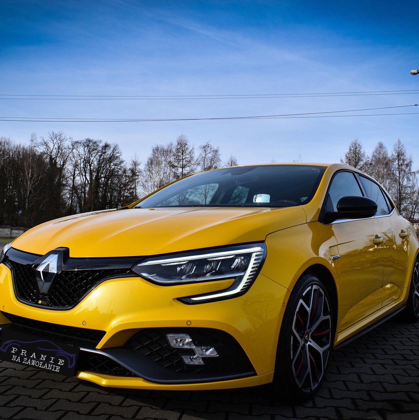 Żółty samochód Renault Megane RS po usłudze czyszczenia, zaparkowany na kostce brukowej, z widocznym logo firmy 'Pranie na Zawołanie' na przednim zderzaku.