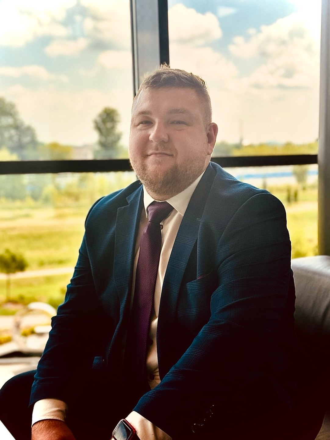 Portret mężczyzny w ciemnym garniturze i bordowym krawacie, siedzącego na tle okna z widokiem na zielony krajobraz.