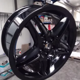 Błyszcząca, czarna felga aluminiowa zawieszona na haku w lakierni proszkowej, widoczne odbicia światła na świeżo pomalowanej powierzchni.