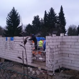 Wznoszenie ścian parteru z białych bloczków betonu komórkowego na budowie domu. Dwóch robotników na rusztowaniu, jeden w kamizelce odblaskowej, muruje ściany. W tle budynek z zielonym dachem i drzewa.