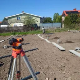 Poziomowanie terenu pod fundament ogrodzenia, widoczny niwelator optyczny na statywie, betonowe bloczki i podkłady ułożone w rzędach, pracownik w oddali.