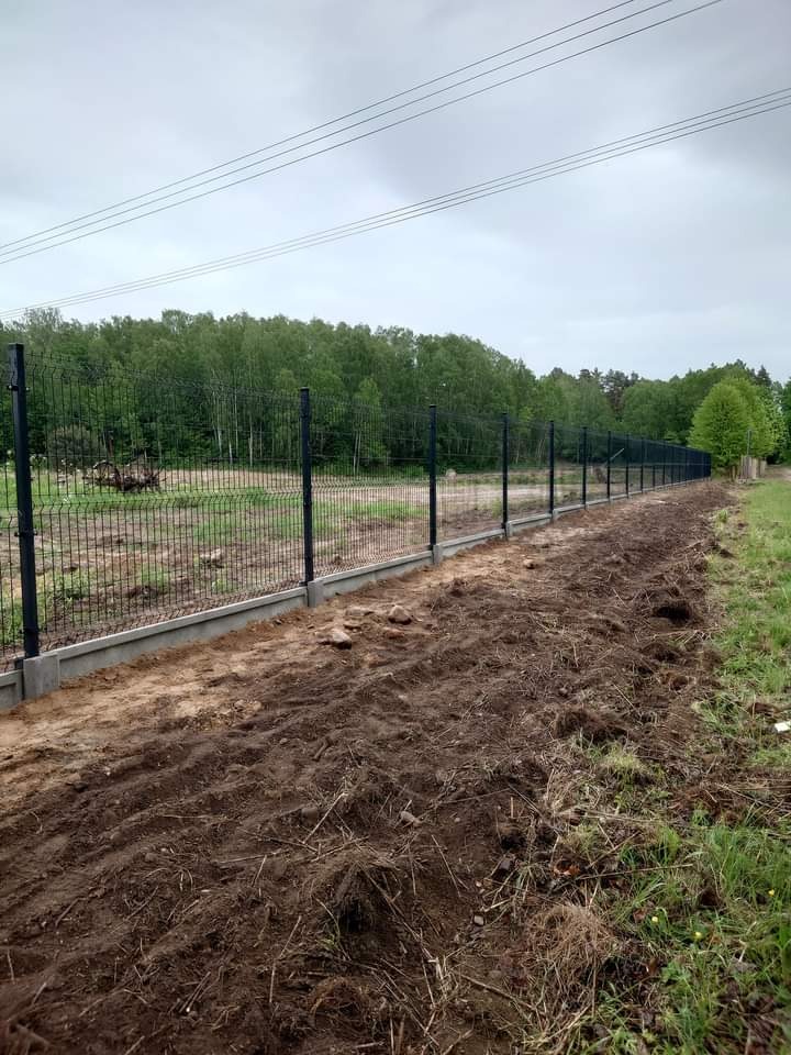 Długie, czarne ogrodzenie panelowe z podmurówką betonową, ciągnące się wzdłuż granicy działki, świeżo po montażu, z widocznymi śladami po pracach ziemnych.
