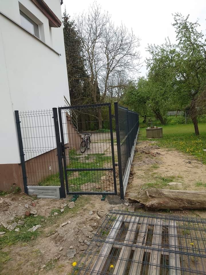 Ciemnoszare ogrodzenie z paneli siatkowych z furtką przy budynku, widoczny rower w tle, na pierwszym planie paleta z siatki ogrodzeniowej.