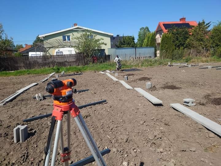 Poziomowanie terenu pod fundament ogrodzenia, widoczny niwelator optyczny na statywie, betonowe bloczki i podkłady ułożone w rzędach, pracownik w oddali.