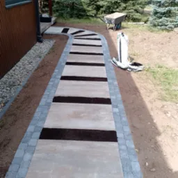 Kaskadowa ścieżka z płyt betonowych z obrzeżem z kostki brukowej, wypełniona ziemią, prowadząca przez ogród z iglakami. W tle taczka i worek ziemi.