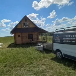 Budowa drewnianego domu z ciemnym dachem w miejscowości Cimochy, widoczne rusztowania i pracownicy na dachu, na pierwszym planie trawa i samochód dostawczy z logo firmy.