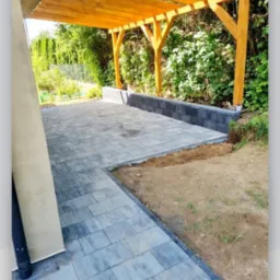 Nowo ułożona, szara kostka brukowa tworząca taras z drewnianą pergolą, widoczne obrzeża i fragment trawnika z ziemią.