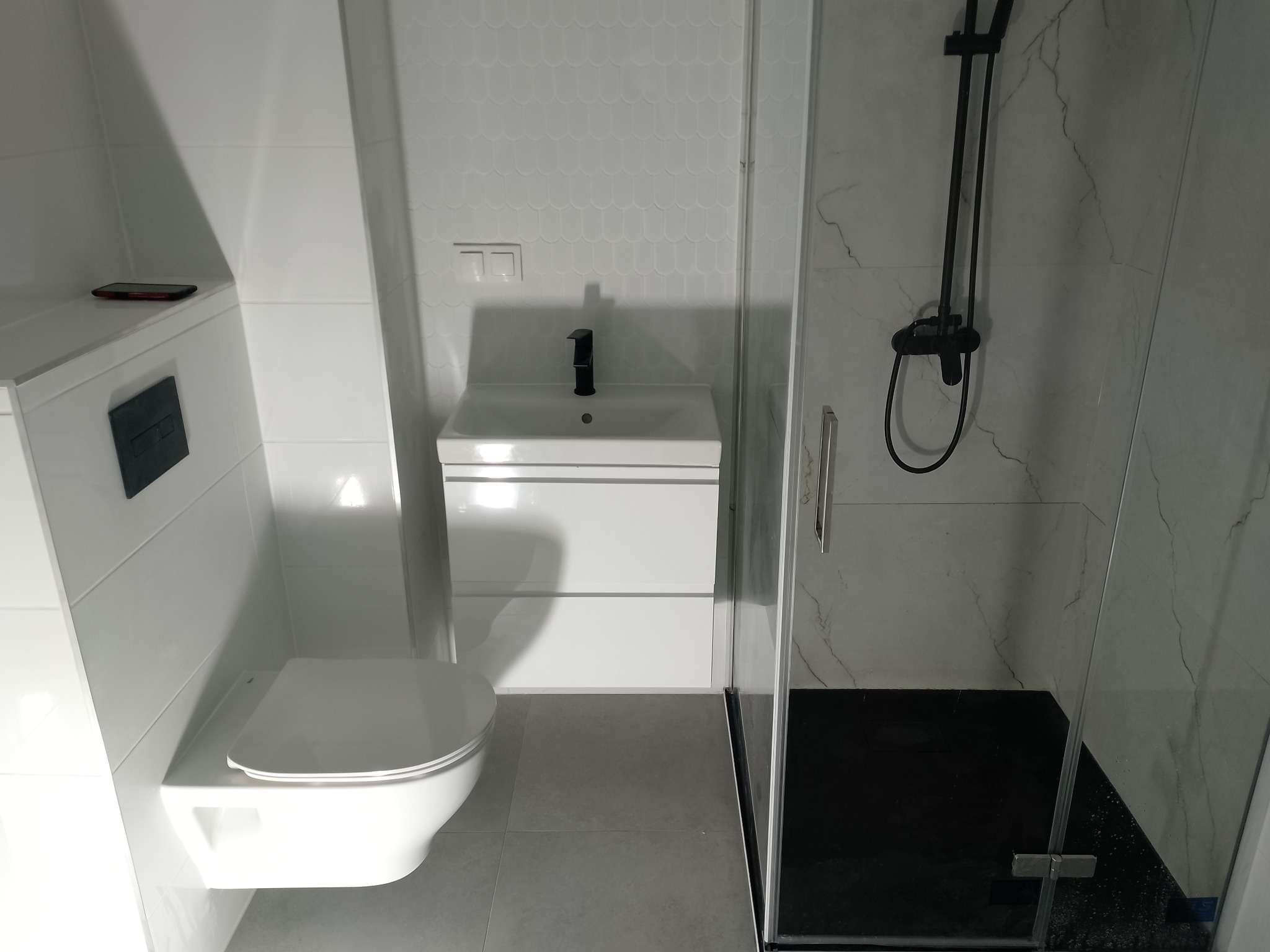 Nowoczesna, jasna łazienka po remoncie: podwieszana toaleta, umywalka z szafką i kabina prysznicowa z czarną armaturą na tle białych płytek.