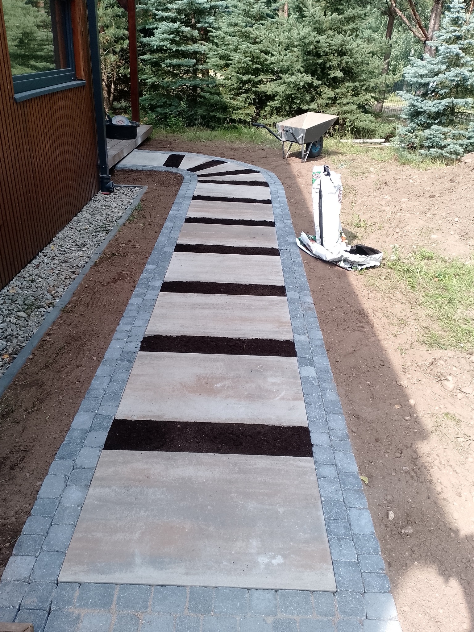 Kaskadowa ścieżka z płyt betonowych z obrzeżem z kostki brukowej, wypełniona ziemią, prowadząca przez ogród z iglakami. W tle taczka i worek ziemi.