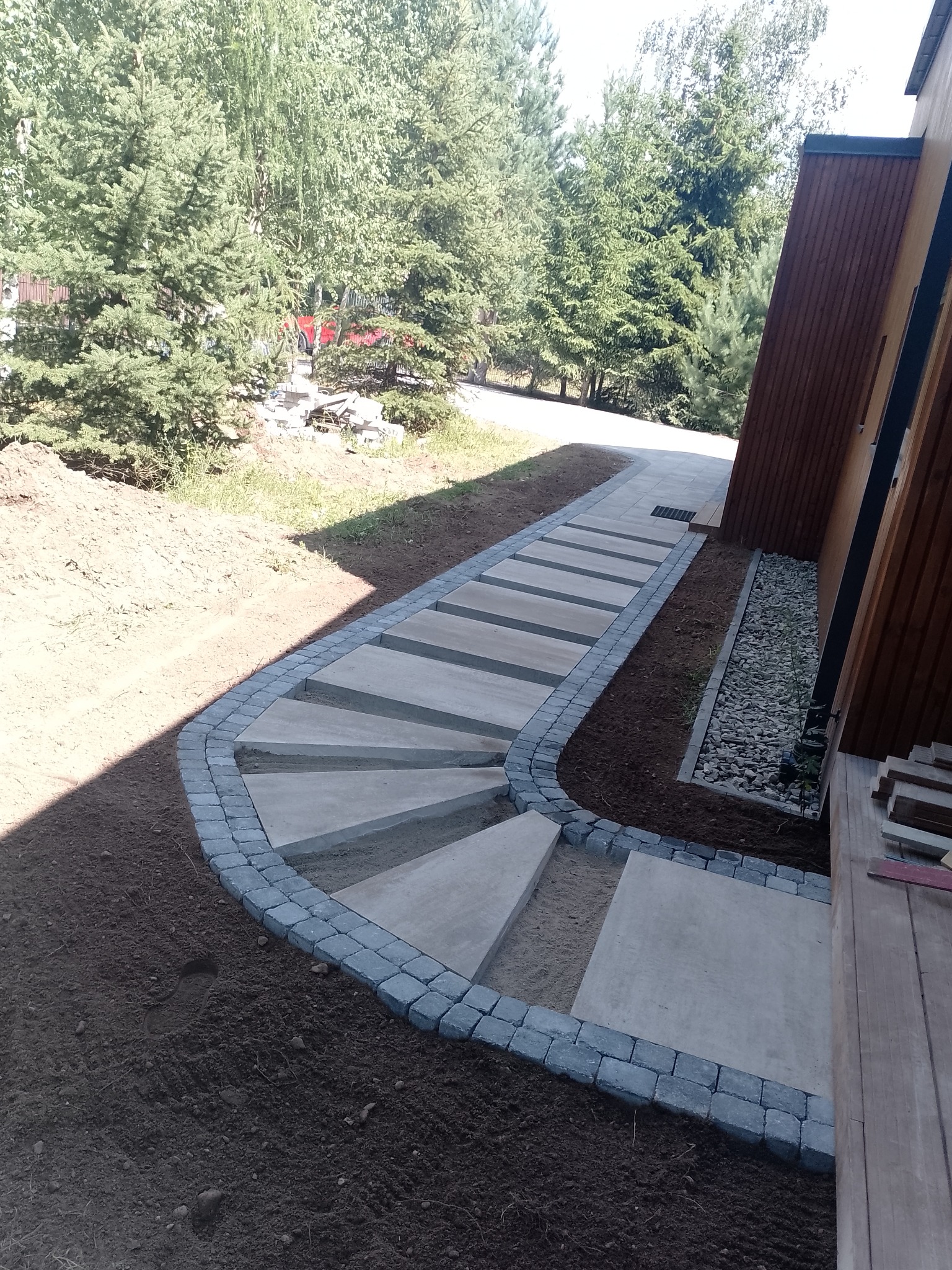 Ścieżka z betonowych płyt obramowana kostką brukową prowadząca od budynku w kierunku ogrodu, widok z góry, świeżo obsypana ziemia po bokach.
