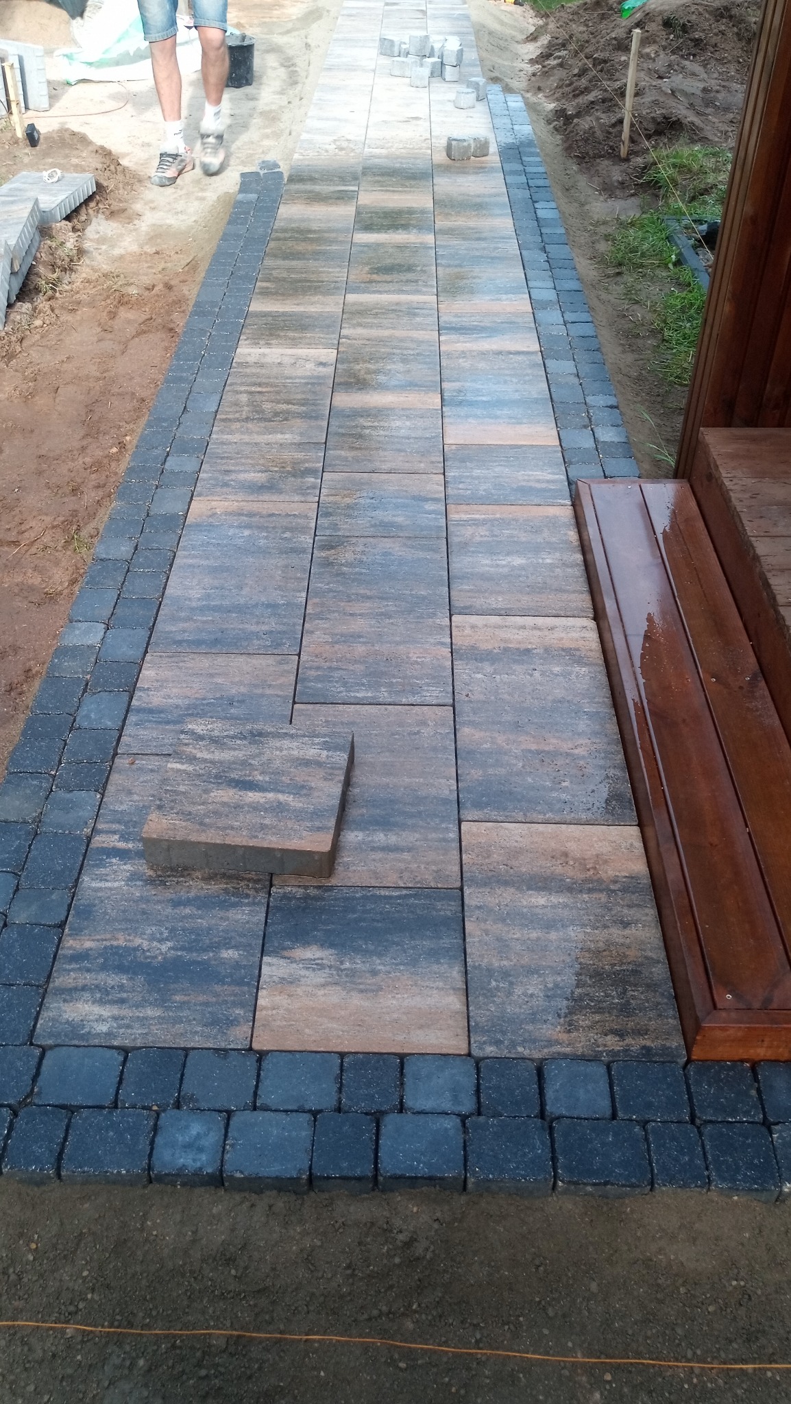 Układanie kostki brukowej w odcieniach szarości i brązu. Częściowo ułożony chodnik z obrzeżami z kostki. Jeden element kostki leży obok, czekając na ułożenie. Widoczne drewniane schody.