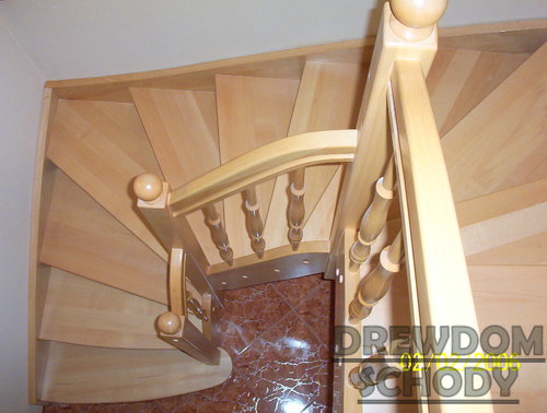 Jasne, drewniane schody spiralne z okrągłymi gałkami na balustradzie, widok z góry na marmurową posadzkę pod schodami, z widocznym znakiem wodnym firmy na dole.