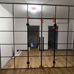 Perfekt Glass - Szklana ściana działowa z czarnymi profilami, widok na wnętrze z drewnianą podłogą. Pomarańczowe elementy montażowe na szkle. Widoczne drzwi dwuskrzydłowe.