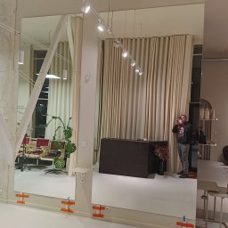 Perfekt Glass - Duże lustra w salonie fryzjerskim, zamontowane na ścianie za pomocą pomarańczowej taśmy. W tle widoczne stanowiska fryzjerskie i osoba robiąca zdjęcie.