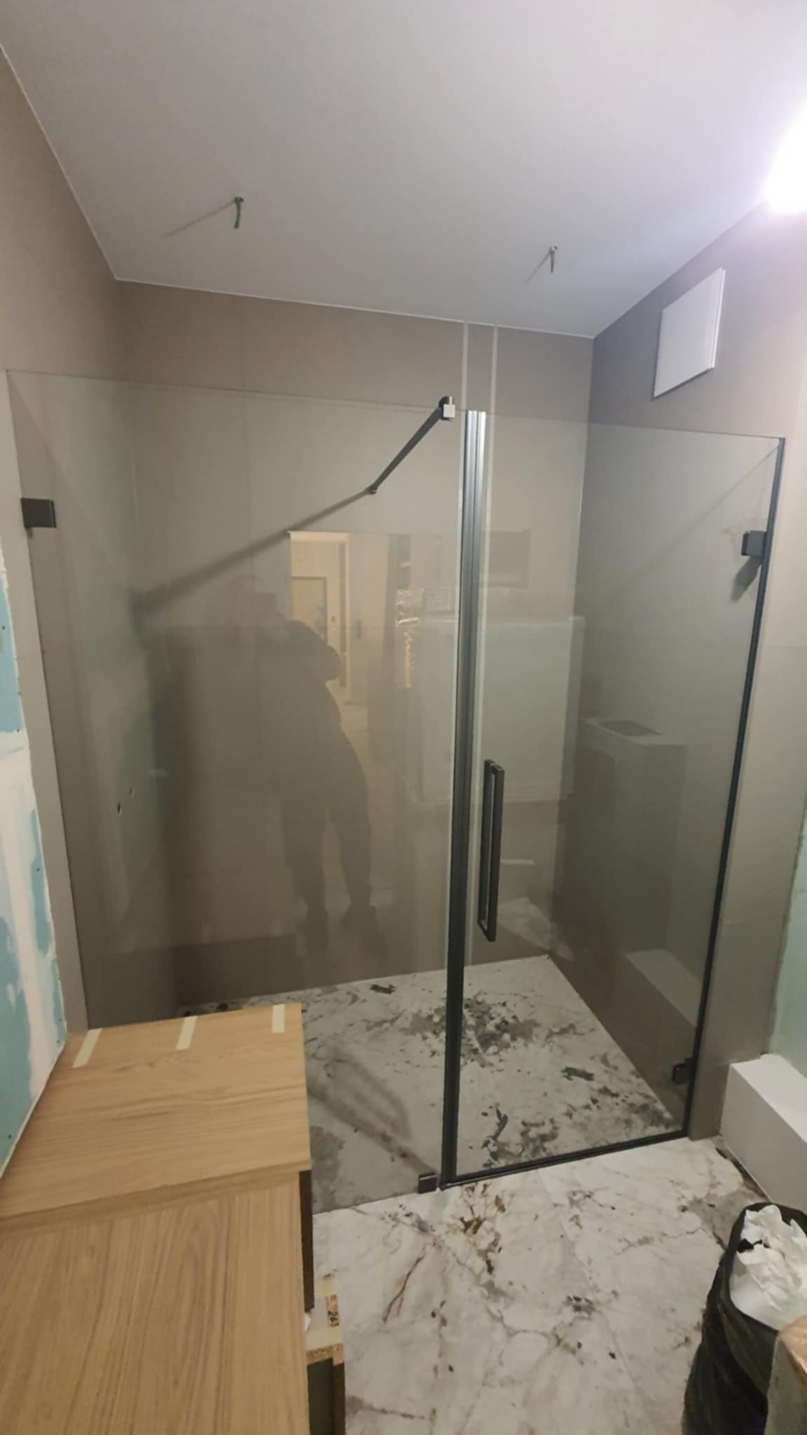 Szklana kabina prysznicowa z czarnymi okuciami w łazience w trakcie remontu. Widoczne niedociągnięcia na podłodze i meble łazienkowe w surowym stanie.
