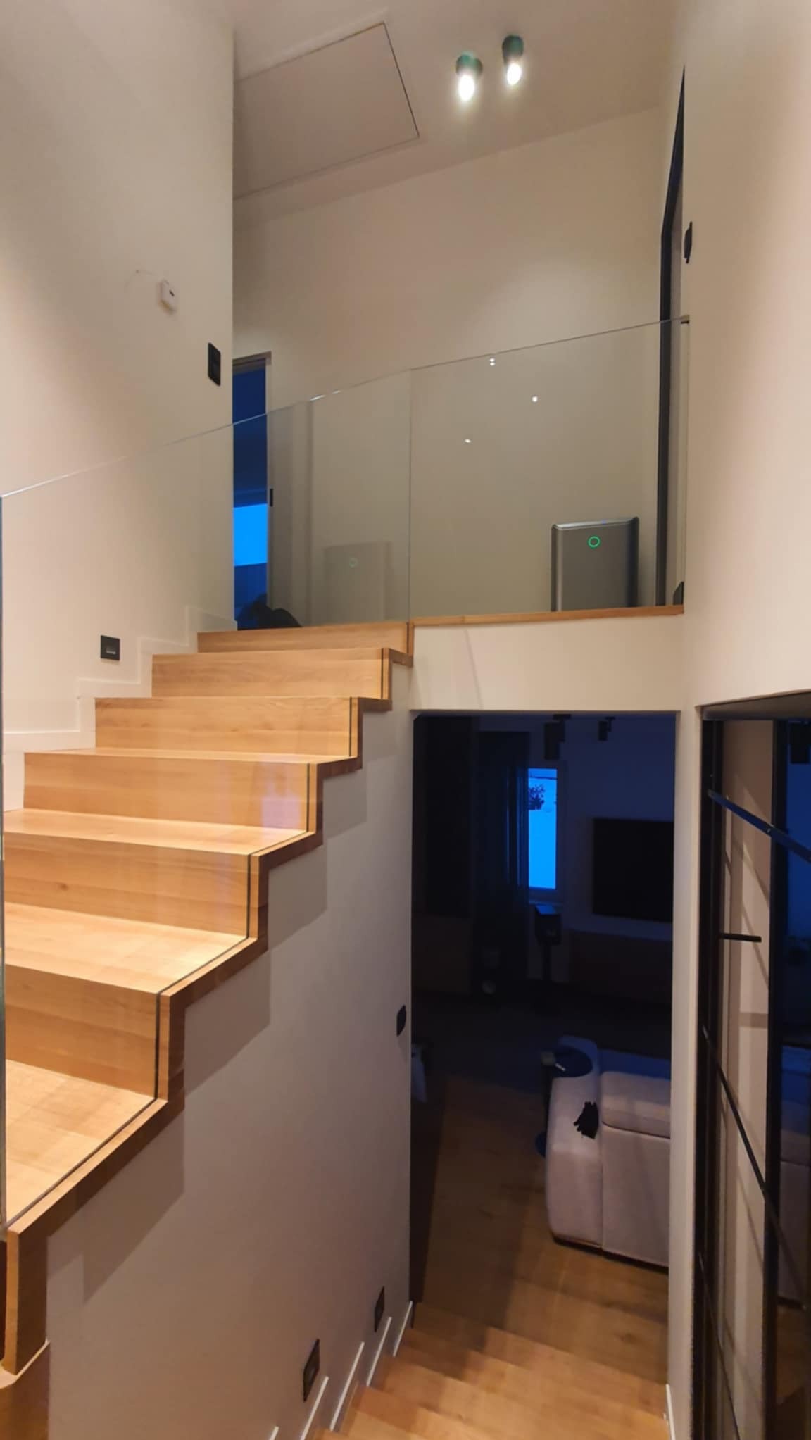 Nowoczesne schody drewniane z przezroczystą, minimalistyczną osłoną. Jasne drewno kontrastuje z bielą ścian. Widoczne oświetlenie i fragment wnętrza domu.