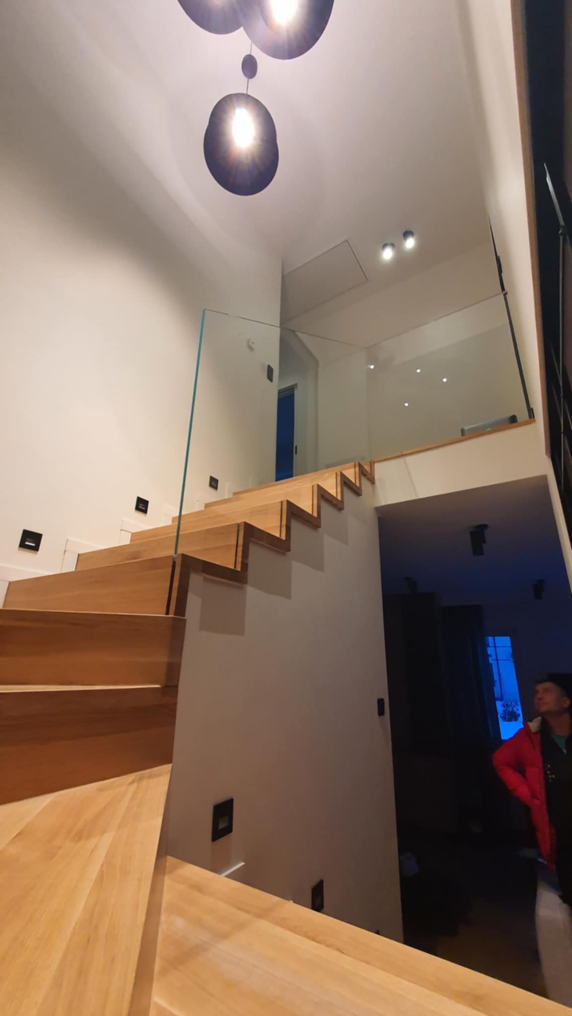 Nowoczesne schody drewniane z przezroczystą, minimalistyczną balustradą szklaną, oświetlone lampami sufitowymi. Widok z dołu, podkreślający design.