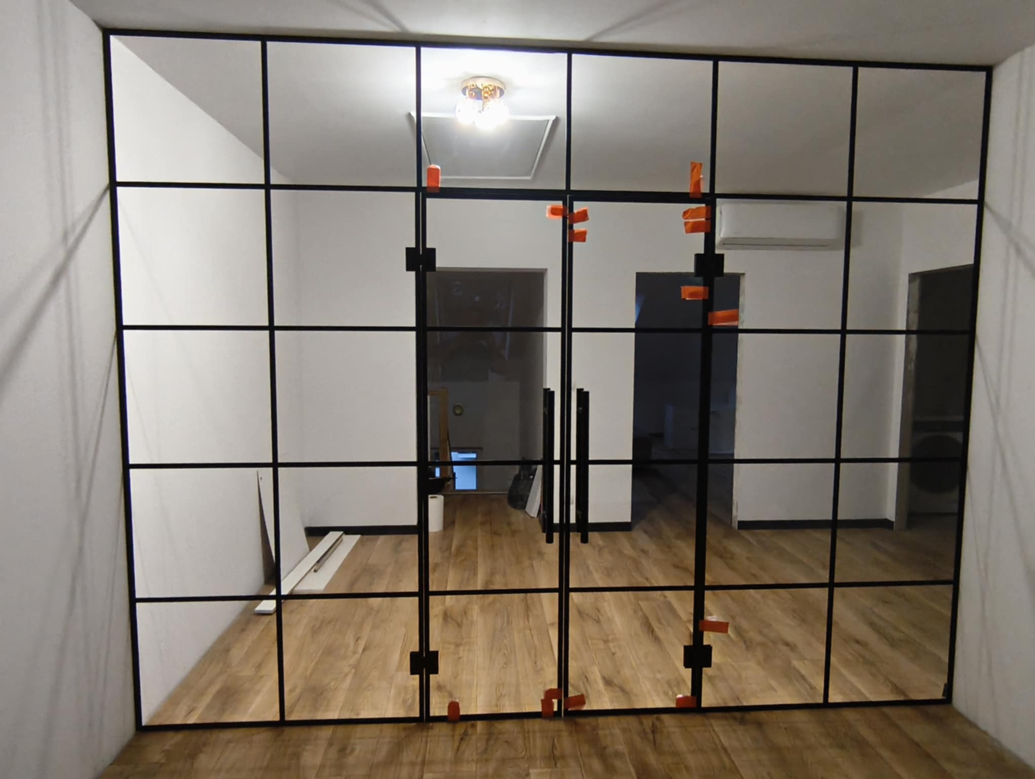 Szklana ściana działowa z czarnymi profilami, widok na wnętrze z drewnianą podłogą. Pomarańczowe elementy montażowe na szkle. Widoczne drzwi dwuskrzydłowe.