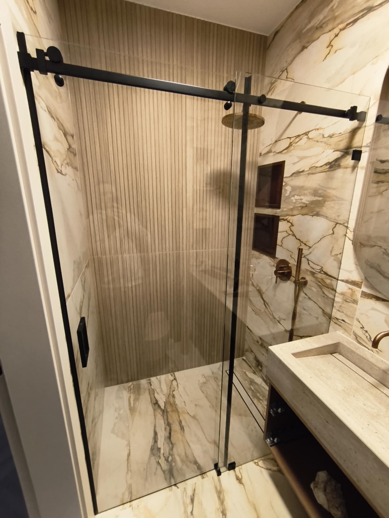 Nowoczesna kabina prysznicowa z przesuwnymi drzwiami, czarne profile, marmurowe wykończenie ścian i podłogi, widoczna armatura w kolorze mosiądzu.