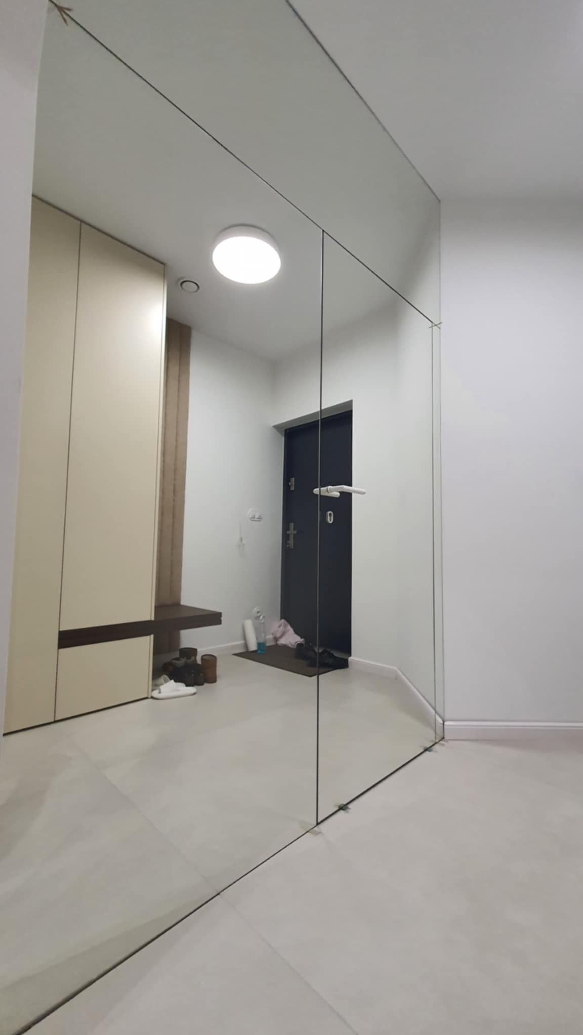 Nowoczesna tafla lustra w przedpokoju, tworząca iluzję przestrzeni. Minimalistyczny design, jasne kolory, odbicie szafy i drzwi wejściowych.