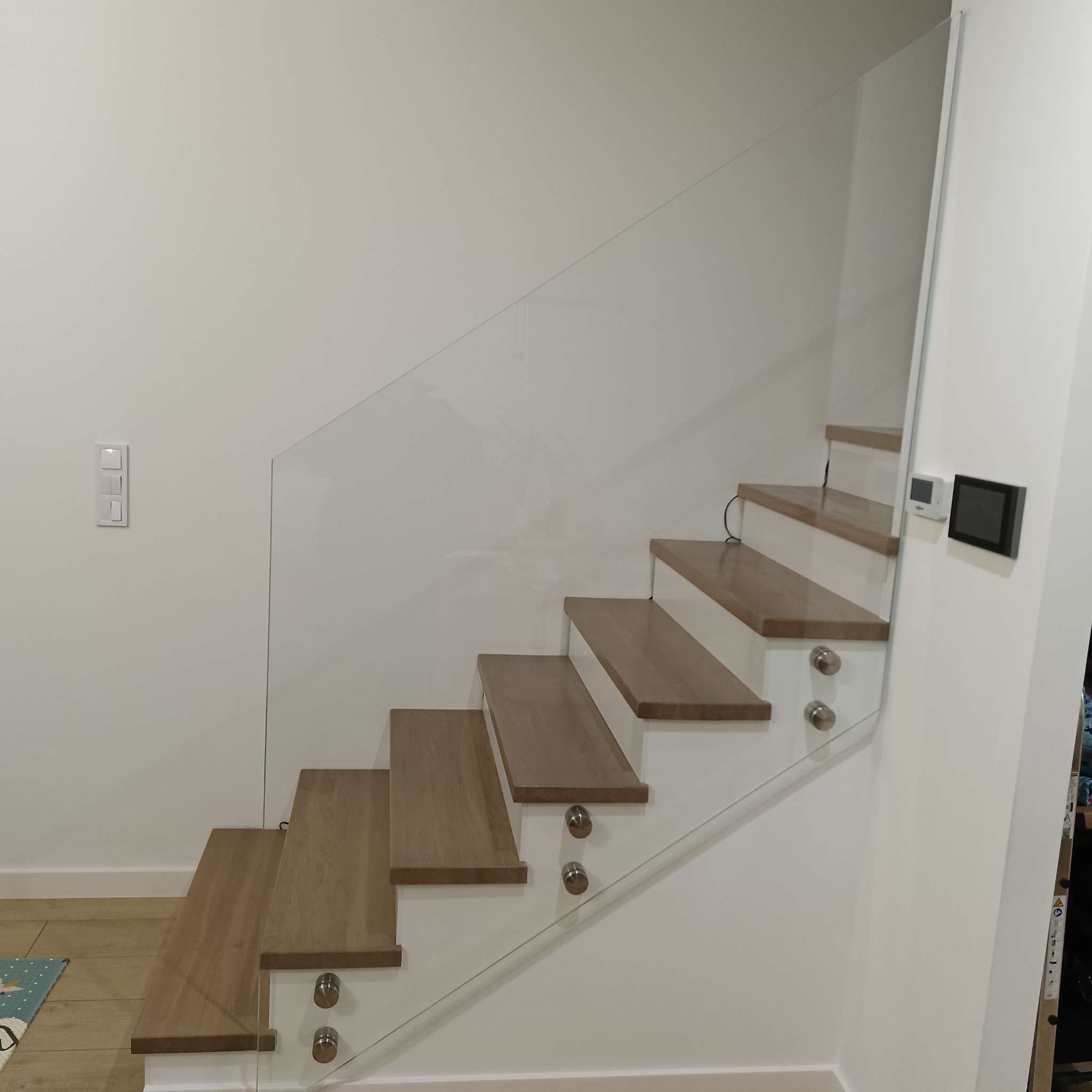 Nowoczesne schody z drewnianymi stopniami i szklaną balustradą, mocowaną na metalowych uchwytach. Minimalistyczny design, jasne wnętrze.