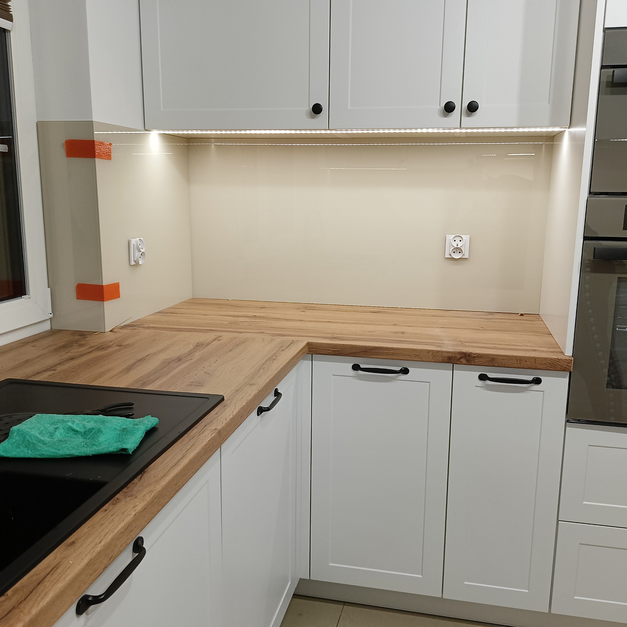 Jasna kuchnia po montażu paneli szklanych nad blatem, z widocznymi gniazdkami i oświetleniem LED. Zabezpieczenia narożników pomarańczową taśmą.