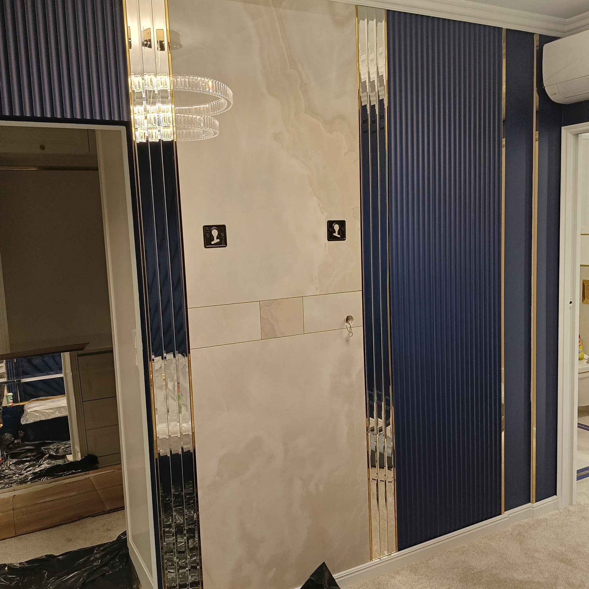 Elegancka ściana z panelami dekoracyjnymi w odcieniach beżu i granatu, z lustrzanymi wstawkami i złotymi akcentami. Widoczne elementy oświetlenia i wykończenia wnętrz.