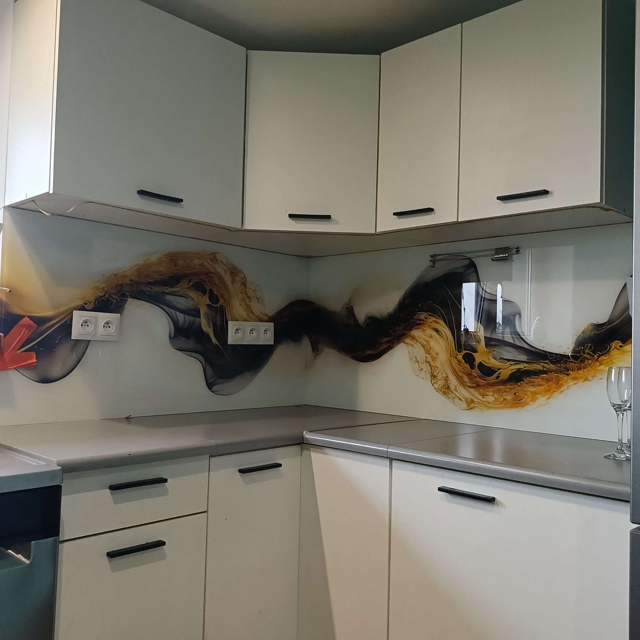 Szklany panel kuchenny z abstrakcyjnym wzorem w odcieniach złota i szarości, zamontowany nad blatem i między szafkami. Widoczne gniazdka elektryczne i fragment szklanki.