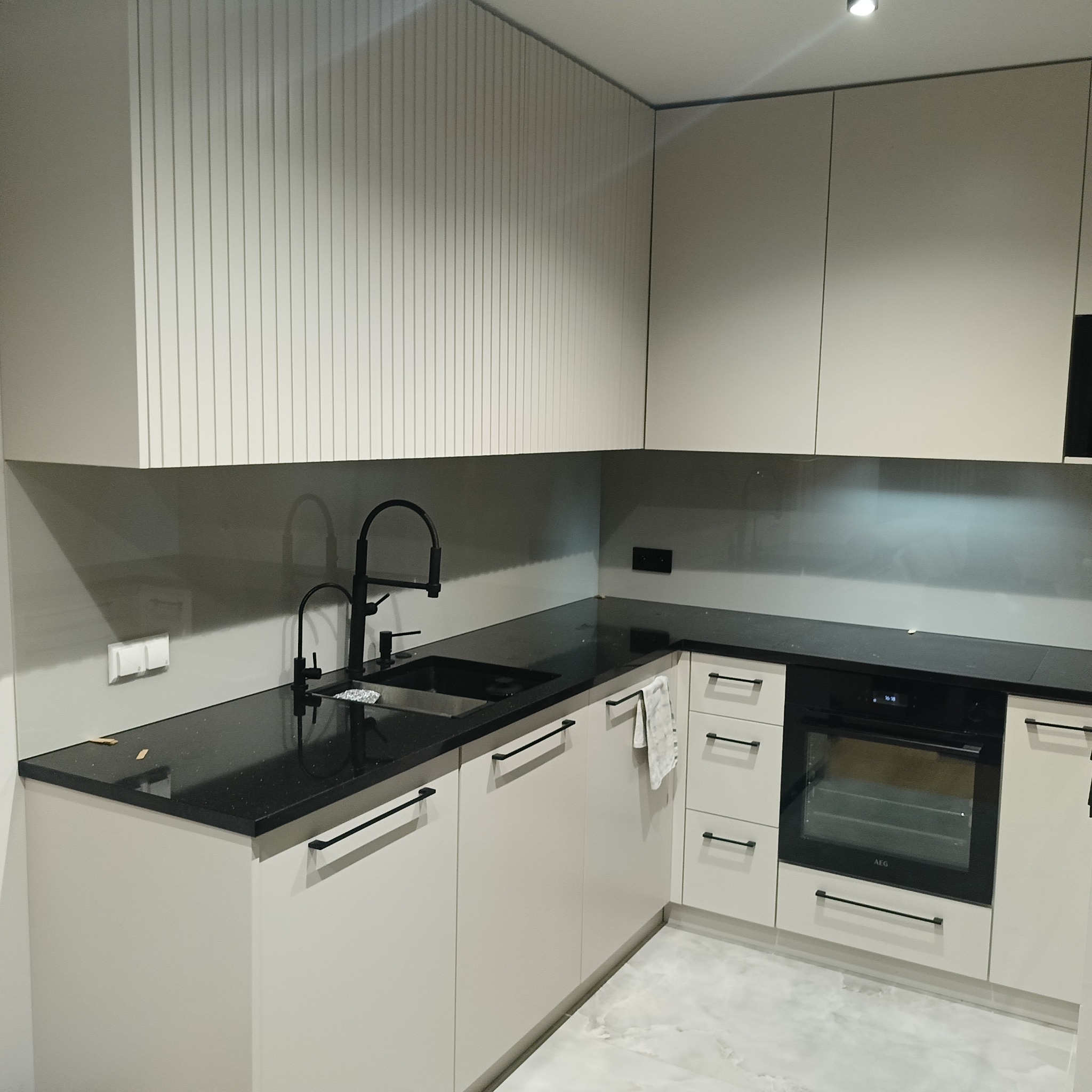 Nowoczesna kuchnia z czarnym blatem i szarym panelem szklanym nad nim. Minimalistyczne szafki w jasnym odcieniu, czarna armatura i piekarnik AEG.