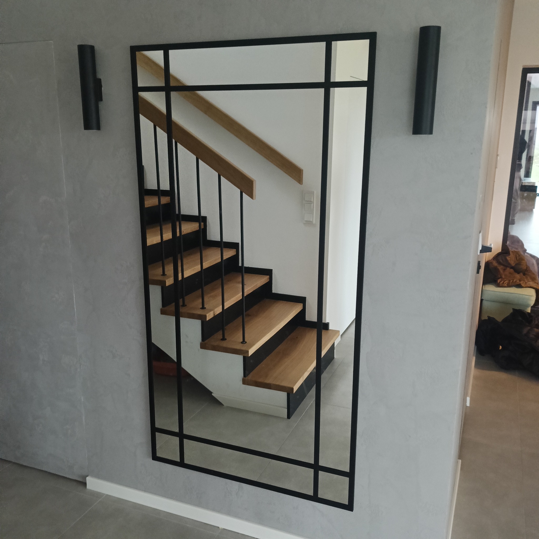Nowoczesne lustro w czarnej ramie, odbijające schody z drewnianymi stopniami i czarną balustradą. Dwa czarne kinkiety na szarej ścianie. Minimalistyczny design.