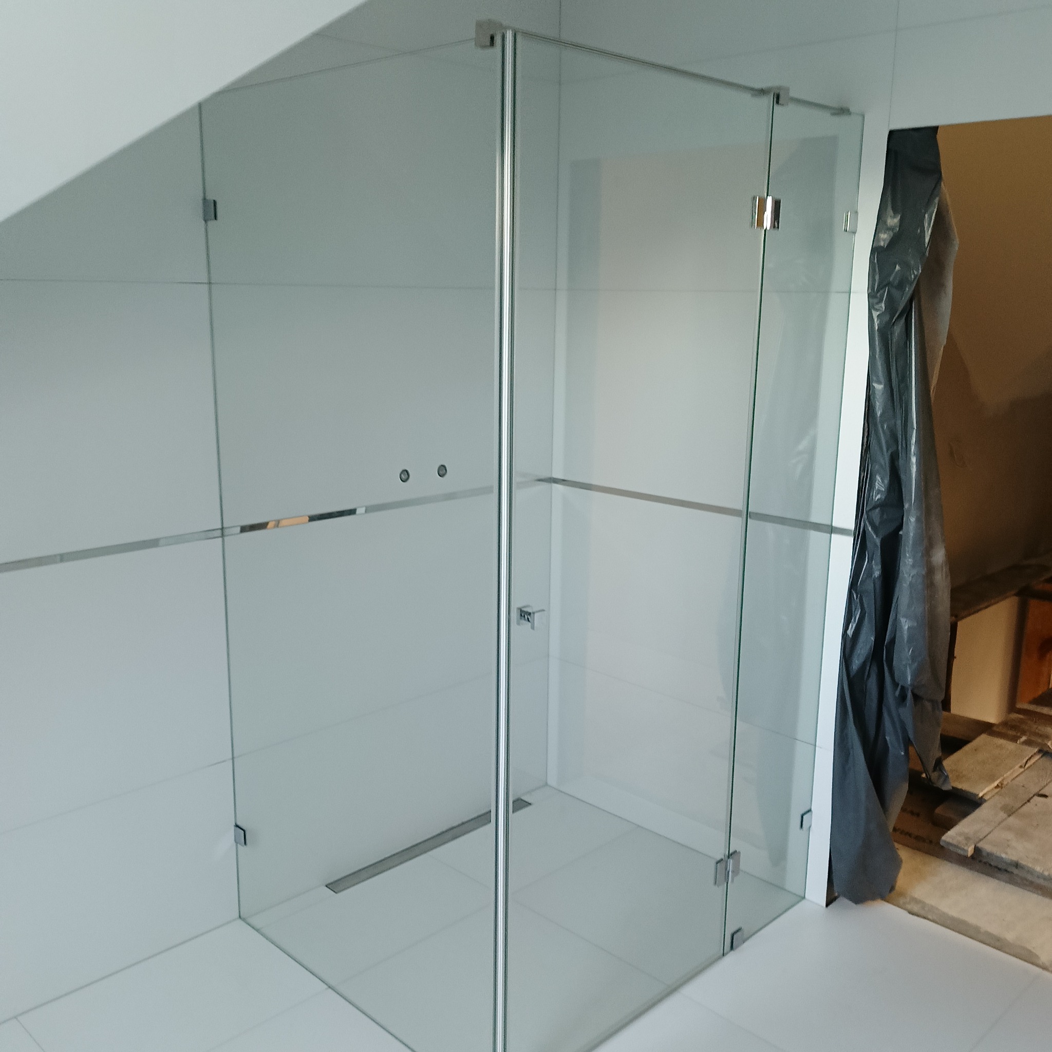 Szklana kabina prysznicowa z chromowanymi okuciami w nowoczesnej łazience wyłożonej białymi płytkami. Widoczny odpływ liniowy i fragment remontowanego pomieszczenia obok.