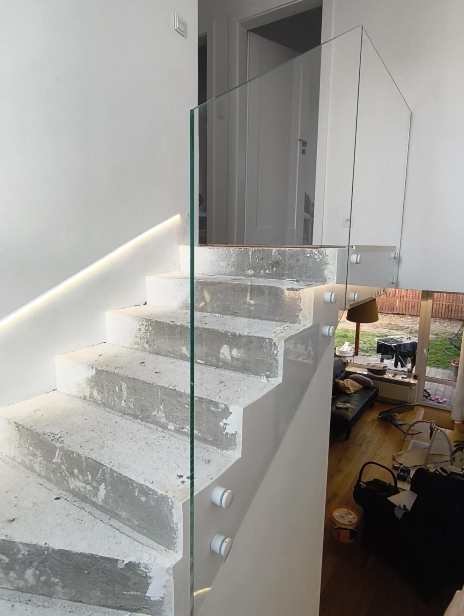 Betonowe schody z zamontowaną szklaną taflą. Widoczne białe mocowania. W tle jasne wnętrze domu z drewnianą podłogą i meblami. Oświetlenie LED na ścianie.