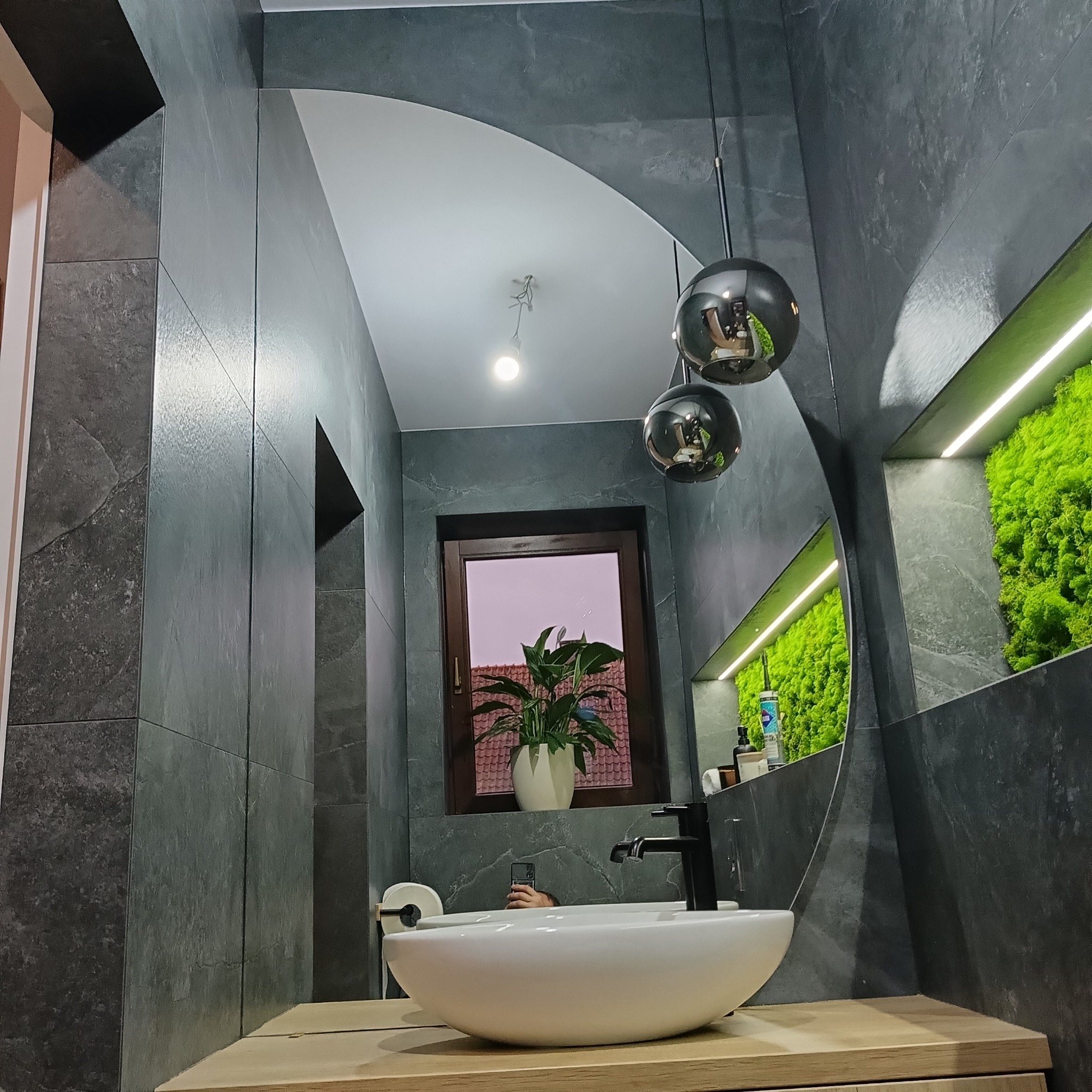 Nowoczesna łazienka z umywalką na drewnianym blacie, szarą okładziną ścian, lustrem o nietypowym kształcie i dekoracyjnym mchem w podświetlanej wnęce.