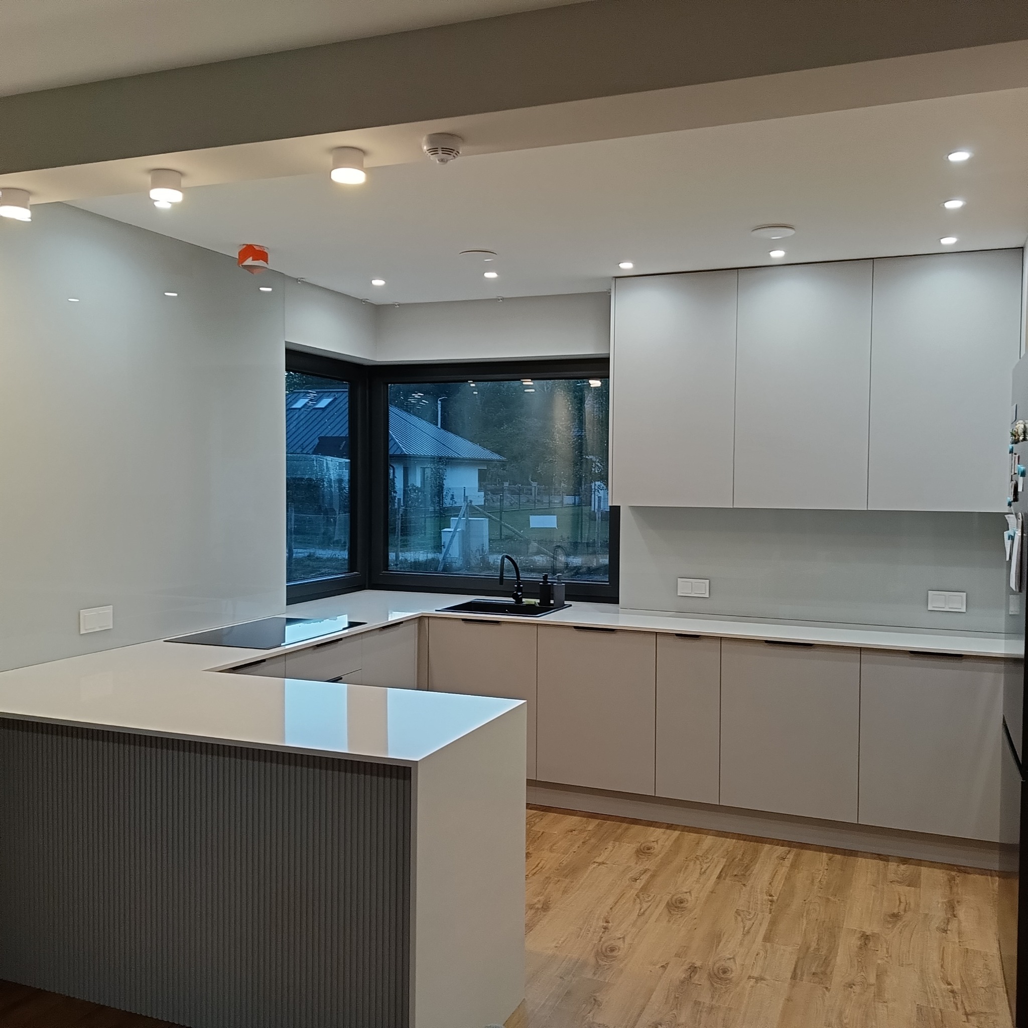 Nowoczesna kuchnia z szarymi szafkami i blatem, szklany panel nad blatem, czarne okno z widokiem na zewnątrz, drewniana podłoga i oświetlenie sufitowe.