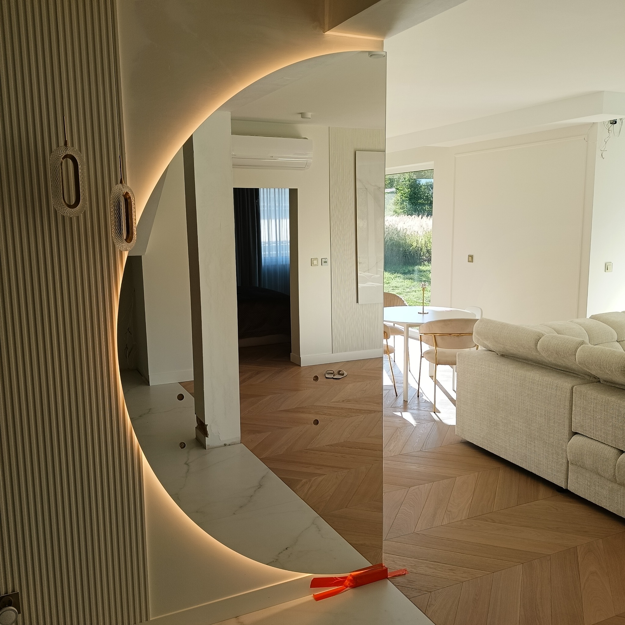 Nowoczesne lustro z podświetleniem LED w kształcie półkola, wkomponowane w ścianę z dekoracyjnymi panelami. Widok na salon z parkietem w jodełkę i jasnymi meblami.