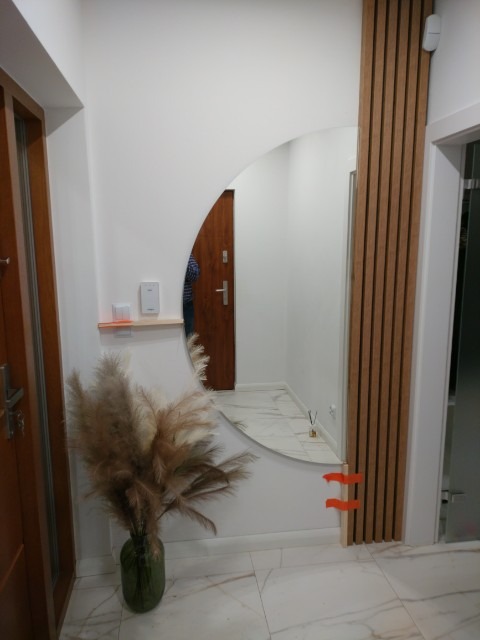 Półokrągłe lustro na ścianie w przedpokoju, z widocznymi elementami dekoracyjnymi: drewniane lamele, trawa pampasowa w szklanym wazonie, marmurowe płytki podłogowe.