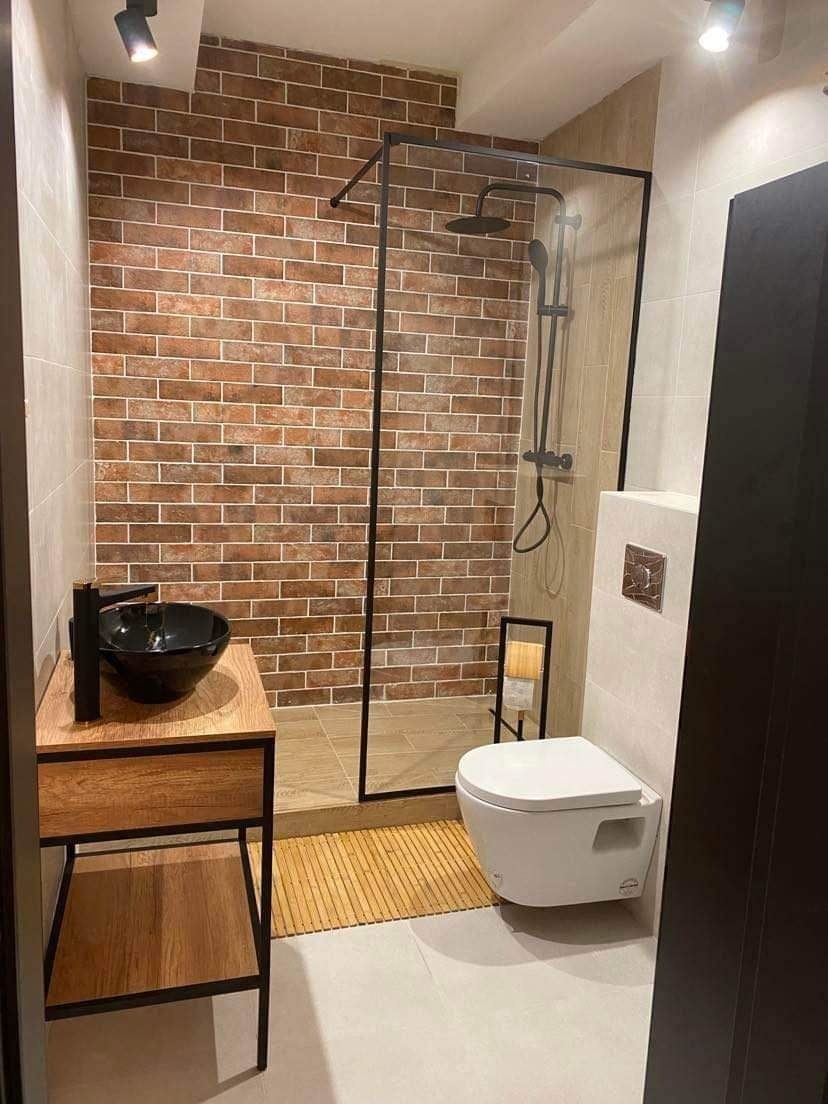 Nowoczesna łazienka z kabiną prysznicową oddzieloną szklaną ścianą z czarną ramą, umywalką nablatową na drewnianej szafce i toaletą podwieszaną.