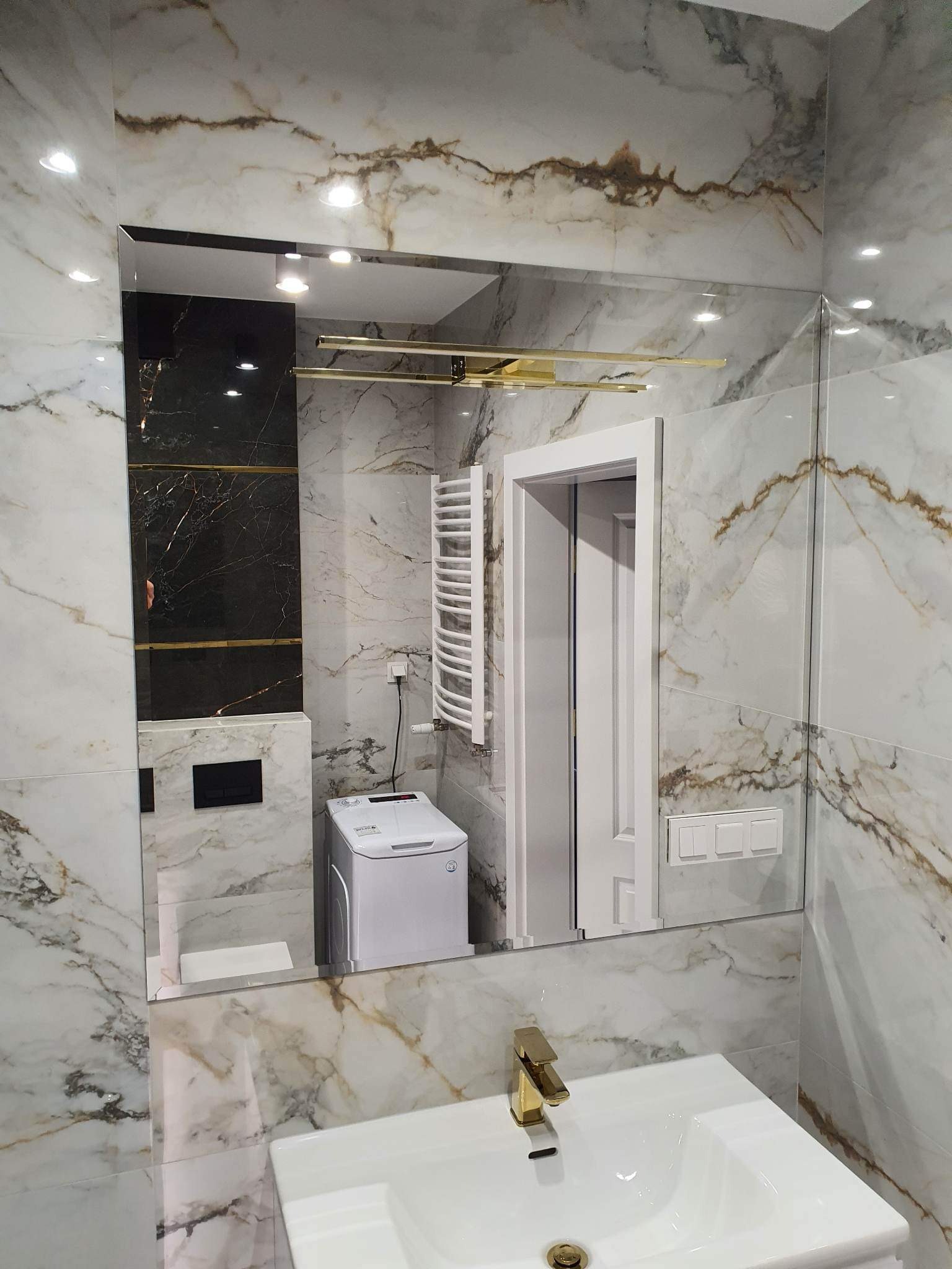Nowoczesna łazienka z lustrem bez ramy, marmurowymi płytkami, złotą armaturą i pralką w tle.