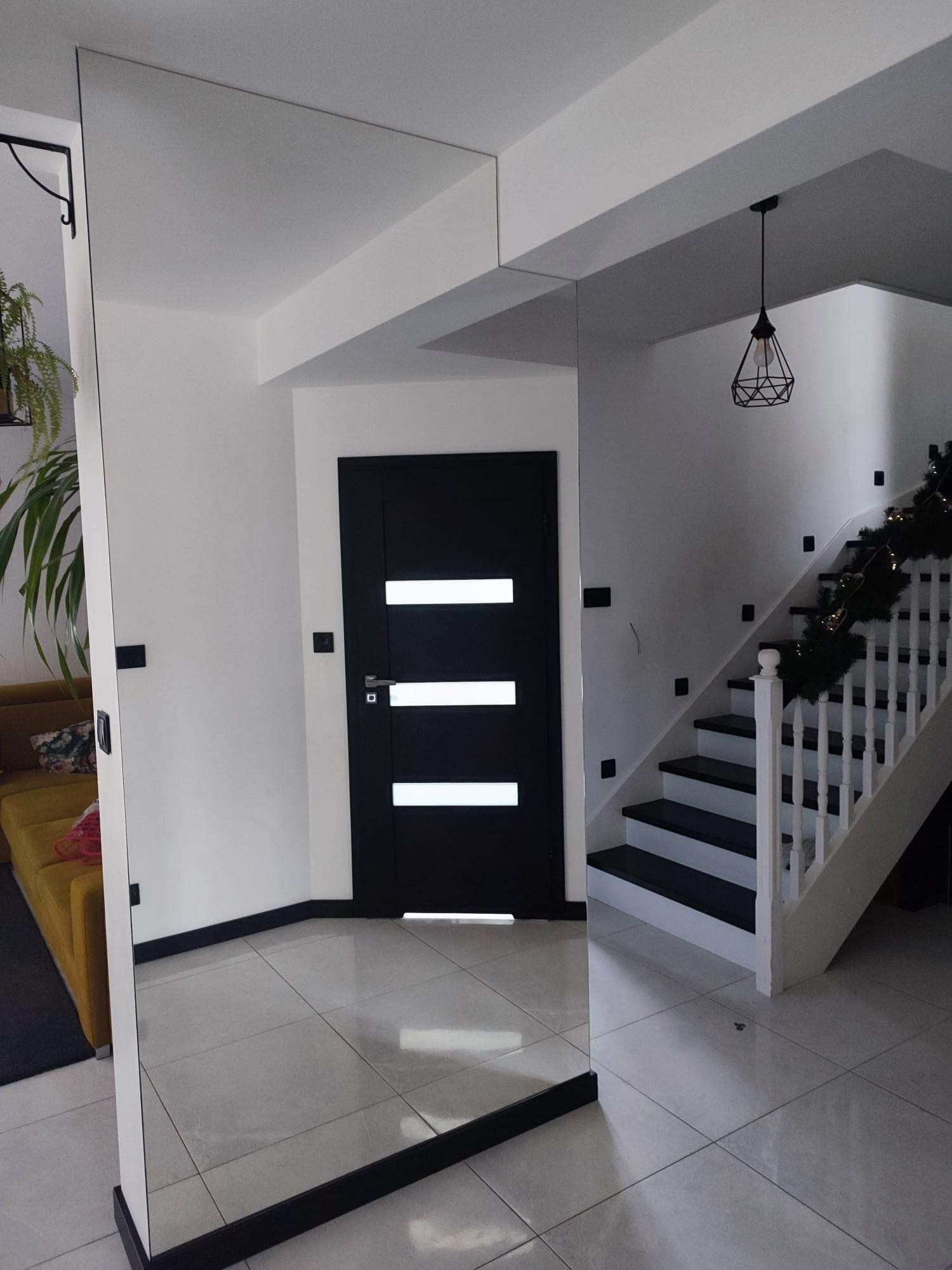 Duże lustro na ścianie w przedpokoju, odbijające fragment sofy, z widocznymi schodami w tle i czarnymi akcentami na drzwiach i schodach.