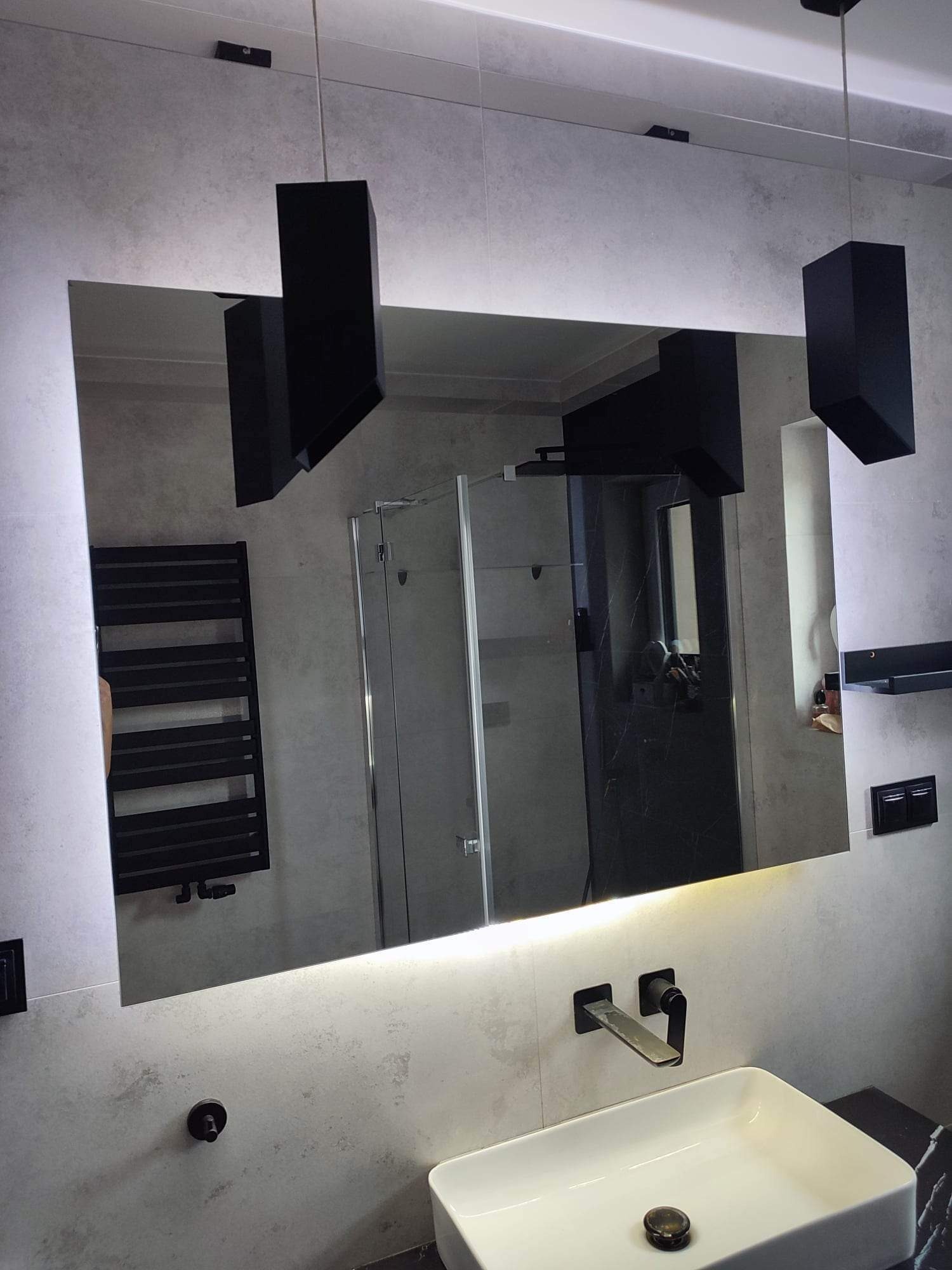 Nowoczesna łazienka z podświetlanym lustrem, wiszącą armaturą i czarnymi elementami dekoracyjnymi, widoczne odbicie kabiny prysznicowej i grzejnika.