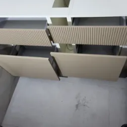 Dwa wysunięte fronty szuflad z beżowego drewna z ryflowanym wzorem, zamontowane w białej szafce, widok z góry na szarą podłogę i marmurowe wykończenie ściany.