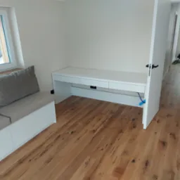 Jasne wnętrze pokoju z drewnianą podłogą, białą zabudową meblową obejmującą biurko z szufladami i siedzisko z poduszkami pod oknem. Widoczny fragment drzwi i dalsza część korytarza.