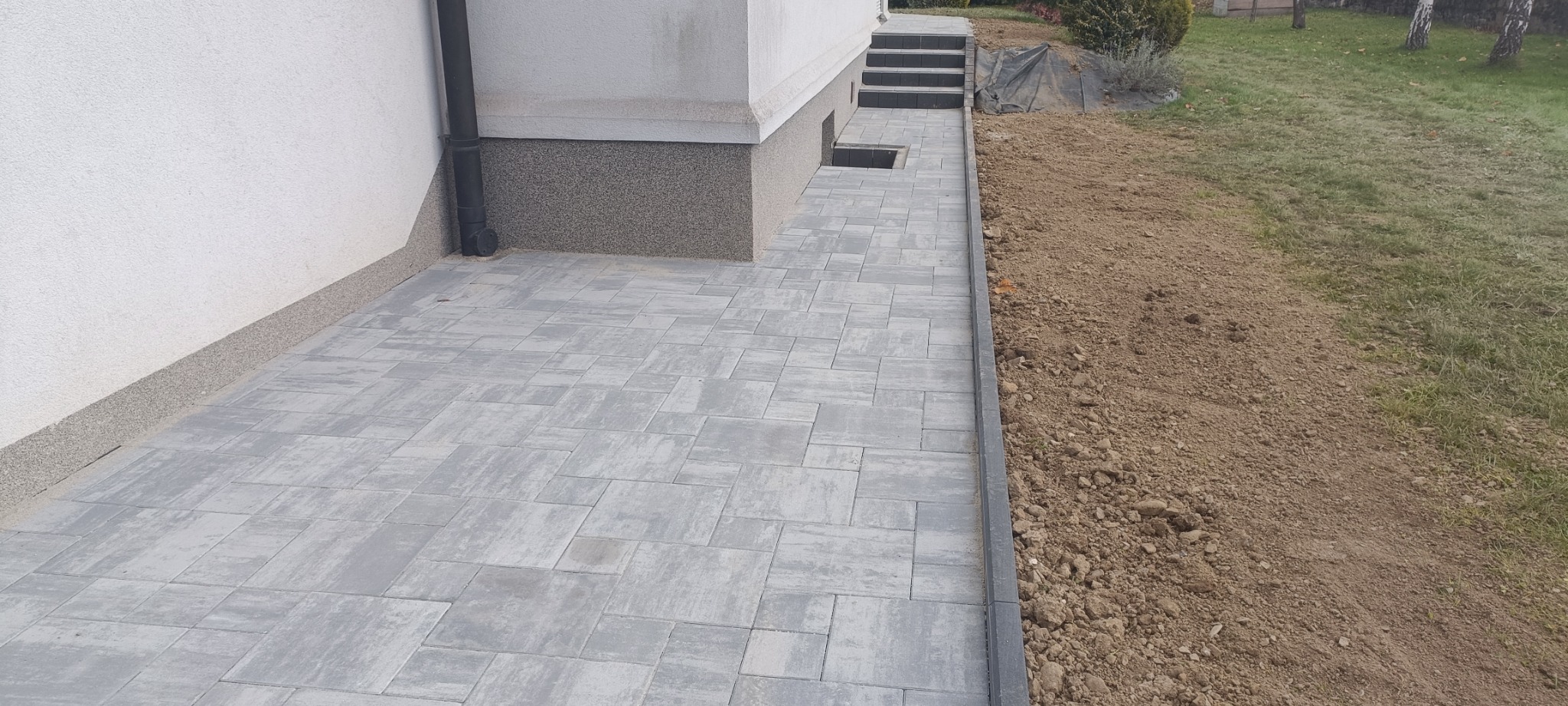 Świeżo ułożona szara kostka brukowa przed budynkiem z widocznym fundamentem i rynną, obok przygotowany teren pod trawnik.