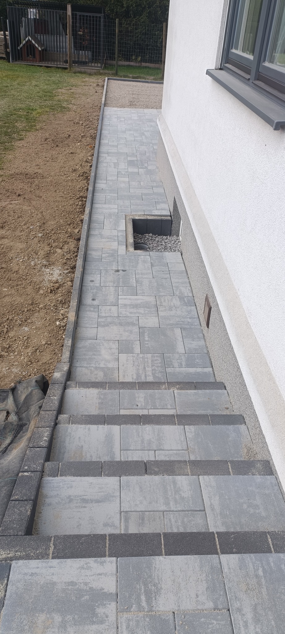 Ułożona szara kostka brukowa prowadząca do budynku, z widocznymi schodami i małym otworem przy ścianie wypełnionym kamieniami.