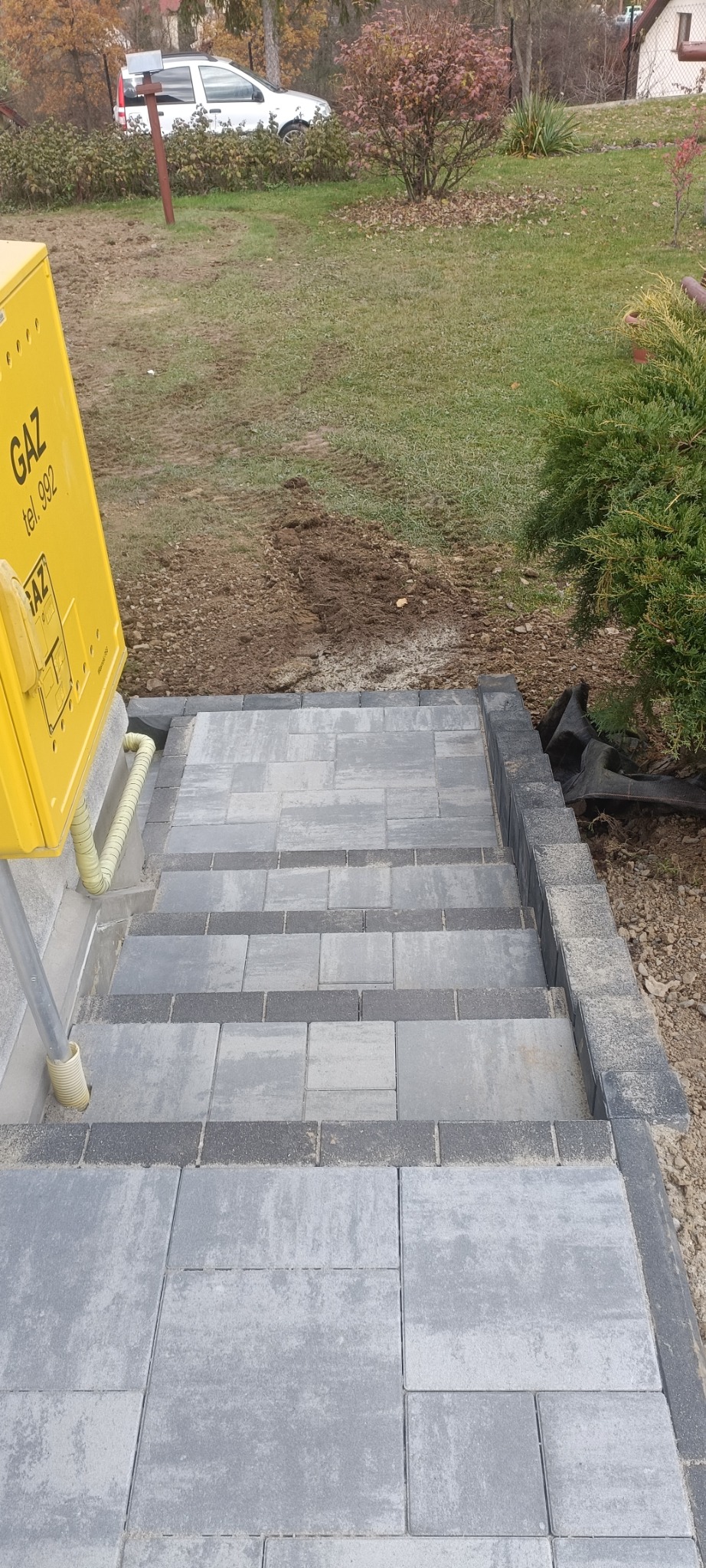Schody z szarej kostki brukowej prowadzące z terenu zielonego do żółtej skrzynki gazowej; widoczne precyzyjne ułożenie i obramowanie ciemniejszą kostką
