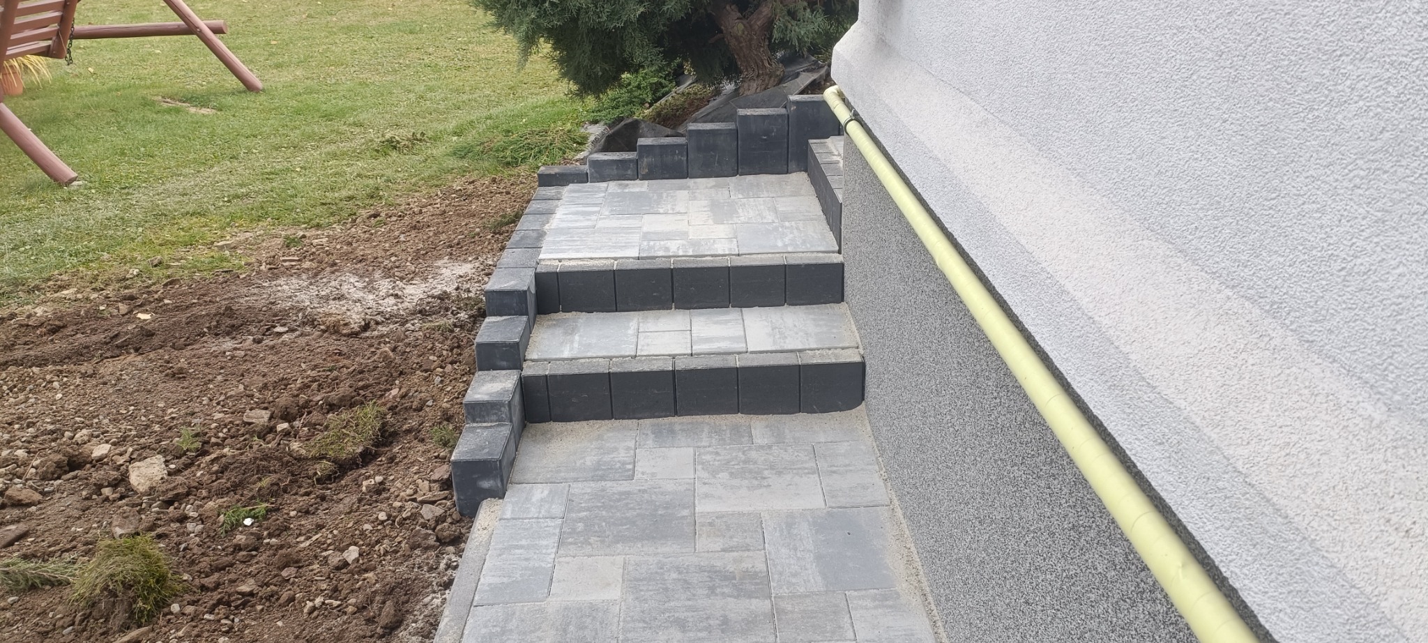 Schody z szarej kostki brukowej prowadzące do budynku z szarą elewacją, obok widoczny trawnik i ziemia.